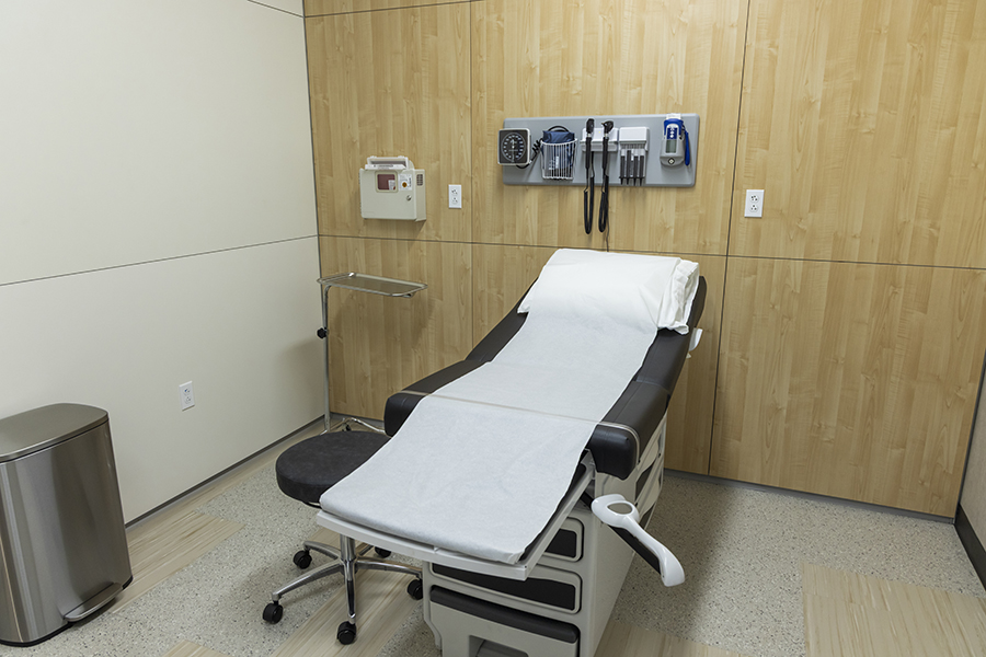 Medical exam room with a black and white examination table covered with a disposable paper sheet. The room has light wood-paneled walls, mounted diagnostic tools such as an otoscope and blood pressure monitor, a rolling stool, and a trash bin in the corner.