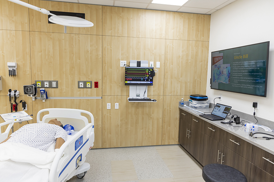 Hospital simulation room with a patient mannequin lying in a hospital bed partially visible on the left. The room has light wood-paneled walls, a wall-mounted monitor displaying medical information, diagnostic equipment, and a countertop with medical supplies and devices along the right side.