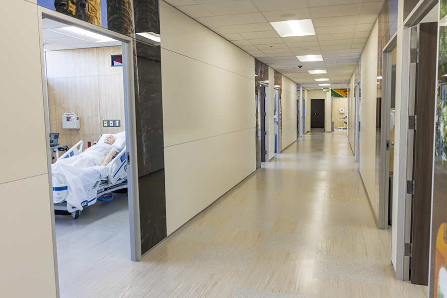 Bright hallway in a medical simulation center with multiple rooms on both sides. One room on the left is open, showing a hospital bed with a patient mannequin covered in white sheets. The corridor has light-colored flooring, white walls, and overhead fluorescent lighting.