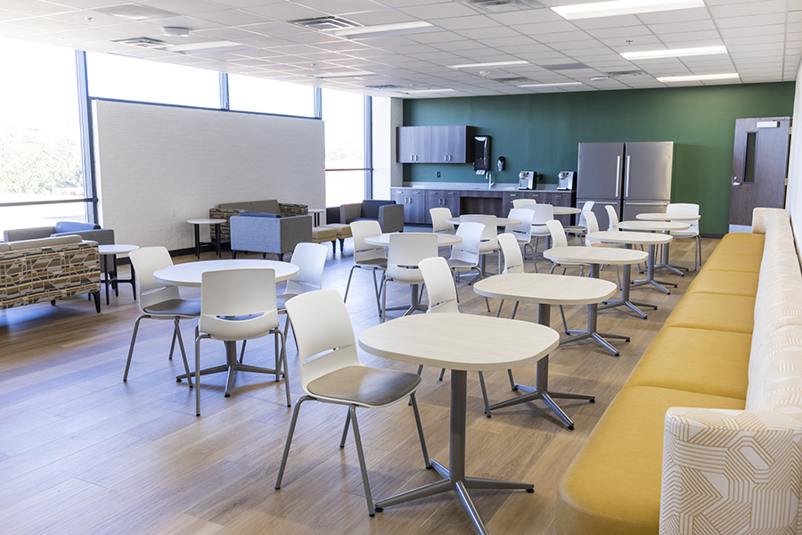 Bright, modern break room with multiple round tables and white chairs arranged in rows. A long yellow cushioned bench runs along one wall. The back of the room features a kitchenette with a refrigerator, microwave, coffee machine, and green accent wall. Large windows provide natural light.