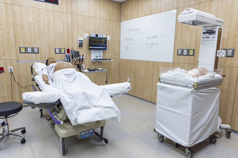 Medical simulation room with two patient mannequins: one lying on a hospital bed covered with white sheets, and another in an infant warmer. The room has light wood-paneled walls, medical equipment mounted on the wall, and a whiteboard in the background.